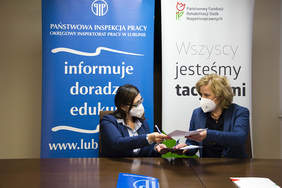 Pokaż zdjęcie: dwie kobiety w niebieskich marynarkach i maseczkach siedzą przy brązowym stole, wymieniają się dokumentami, w tle loga PIP oraz PFRON