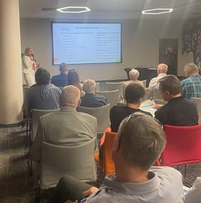 Pokaż zdjęcie: Sala konferencyjna, zdjęcie zrobione z tyłu sali w kierunku prelegenta, kobiety blondynki. Na ekranie slajd z prezentacji.