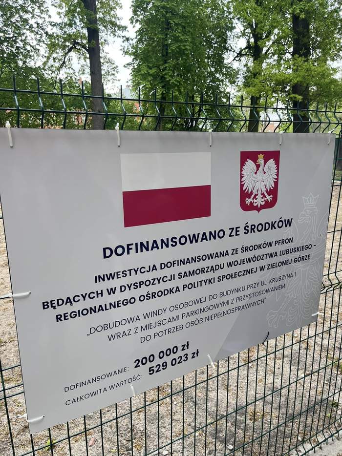 Pokaż zdjęcie: Tablica informacyjna o dofinansowaniu z PFRON windy, powieszona na ogrodzeniu