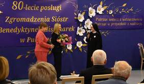 Pokaż zdjęcie: Siostrze zakonnej dwie kobiety składają życzenia, trzymają kwiaty oraz grawerton.