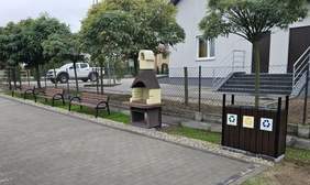 Pokaż zdjęcie: Z przodu znajdują się: śmietnik do segregacji odpadów, grill oraz trzy ławki. W tle można dostrzec: płot, dość niskie, uformowane drzewa, samochód oraz dwa budynki.