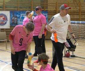 Zawodnicy w różowych koszulkach z numerami oraz mężczyzna w białej koszulce podczas przerwy w rozgrywkach goalball. Jeden z zawodników podaje wodę koledze z drużyny.