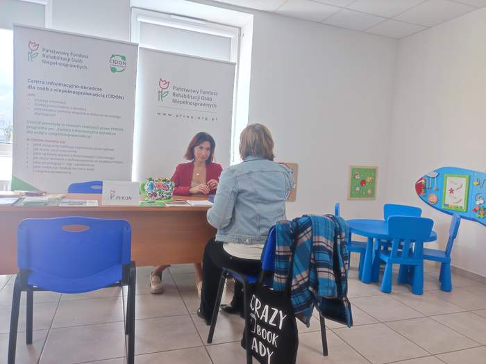 Pokaż zdjęcie: Przys tole na którym leżą ulotki siedzą dwie kobiety, jedna plecami do zdjęcia. W tle rollupy z logo PFRON i CIDON.