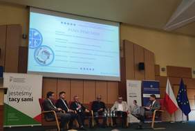 Pokaż zdjęcie: Dyskusyjny panel sześciu mężczyzn siedzących za stołem przed ekranem z napisem „PANEL DYSKUSYJNY” i listą uczestników. Po lewej stronie stoi baner z hasłem „Wszyscy jesteśmy tacy sami” i logo PFRON. W tle po prawej stronie widoczny jest drugi baner z logo PIP oraz flagi Polski i Unii Europejskiej.