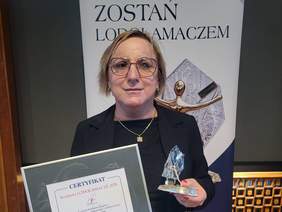 Pokaż zdjęcie: Kobieta w okularach i ciemnej marynarce trzyma oprawiony certyfikat oraz statuetkę Lodołamaczy. W tle widoczny jest napis „Zostań Lodołamaczem” oraz grafika przedstawiająca statuetkę.  