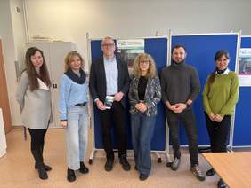 Pokaż zdjęcie: Cztery kobiety i dwóch mężczyzn stoi obok siebie w jasnej sali, pozując do zdjęcia przed niebieskimi tablicami z plakatami informacyjnymi. Jedna z osób trzyma w ręku broszurę. W tle widoczna jest szafa, ściana oraz fragment stołu.