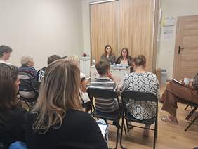 Pokaż zdjęcie: Spotkanie grupy osób w niewielkiej sali. Uczestnicy siedzą na krzesłach ustawionych w rzędach i słuchają dwóch kobiet siedzących przy stole z materiałami informacyjnymi.