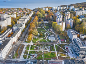 Pokaż zdjęcie: Widok z lotu ptaka na dzielnicę z blokami mieszkalnymi i nowoczesnym parkiem z alejkami, zielenią i infrastrukturą rekreacyjną. W tle morze i portowe statki