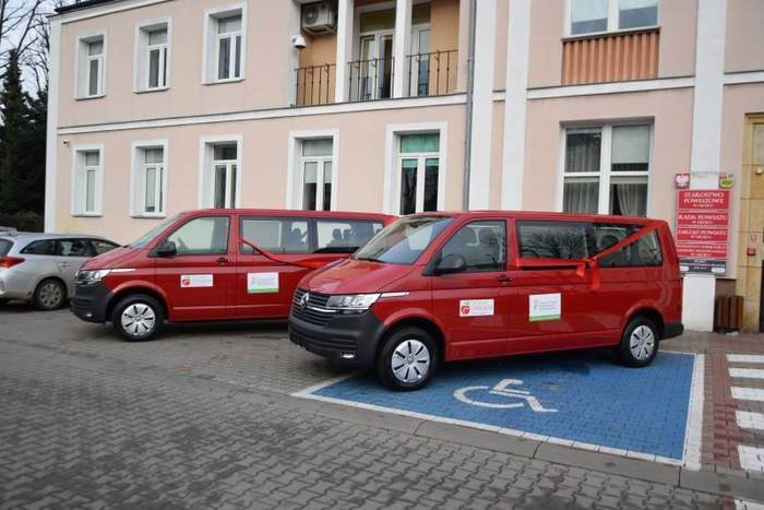 Dwa samochody (busy 9-cio miejscowe) stojące przed budynkiem w którym mieści się siedziba Starostwa Powiatowego w Grójcu. Samochody są koloru wiśniowego, na bokach naklejona m.in. naklejka iformująca o dofinansowaniu zakupu ze środków PFRON.Samochody przepasane czerwoną wstęgą. 
