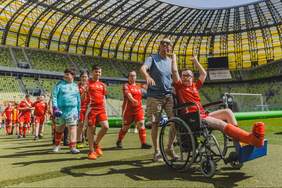 Pokaż zdjęcie: Osoby uczestniczące w turnieju piłkarskim na murawie Polsat Plus Areny Gdańsk. Na pierwszym planie osoba na wózku inwalidzkim w czerwonym stroju sportowym, obok opiekun i grupa zawodników. W tle widoczne trybuny stadionu i boisko.