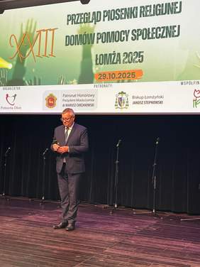 Pokaż zdjęcie: Na środku sceny stoi mężczyzna z mikrofonem, który przemawia do publiczności. Nad nim znajduje się informacja, że jest to XXIII Przegląd Piosenki Religijnej Domów Pomocy Społecznej Łomża 2025, projekt współfinansowany ze środków Państwowego Funduszu Rehabilitacji Osób Niepełnosprawnych.