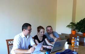 Pokaż zdjęcie: Trzy osoby. Siedzą przy stoliku, na którym stoi laptop i leżą dokumenty.
