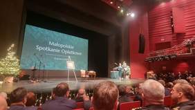 Pokaż zdjęcie: Widok sali Opery Krakowskiej podczas Małopolskiego Spotkania Opłatkowego – na scenie stoją prowadzący przy mównicy, obok choinki i elementy świątecznej dekoracji, w tle ekran z napisem „Małopolskie Spotkanie Opłatkowe, 16 grudnia 2025”, na widowni zgromadzeni uczestnicy wydarzenia.