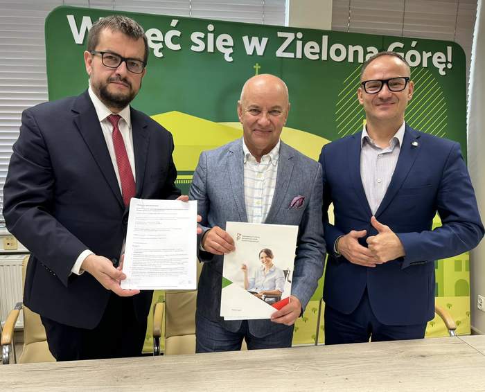 Pokaż zdjęcie: Na tle plakatu promującego Zieloną Górę stoją trzej mężczyźni w garniturach, są uśmiechnięci, dwóch z nich trzyma umowy w ręku