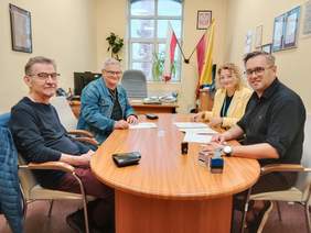 Pokaż zdjęcie: Przy podłużnym stole siedzi kobieta i trzech mężczyzn. Wszyscy patrzą w stronę aparatu. Zawierają umowę. Na stole leżą różne dokumenty. 
