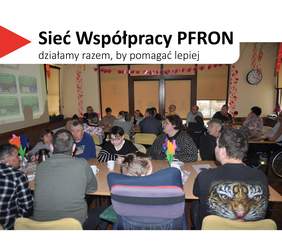 Pokaż zdjęcie: Sala wypełniona dorosłymi uczestnikami siedzącymi przy stołach, zwróconymi w stronę prowadzącego. W lewym górnym rogu widnieje hasło „Sieć Współpracy PFRON – działamy razem, by pomagać lepiej”.