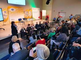 Pokaż zdjęcie: Na scenie siedzą na krzesłach uczestnicy debaty konferencyjnej. Rozmawiają ze sobą. Przysłuchują się im goście na widowni. 