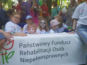 Dzieci z pomalowanymi twarzami oparte o baner z napisem Państwowy Fundusz Rehabilitacji Osób Niepełnosprawnych
