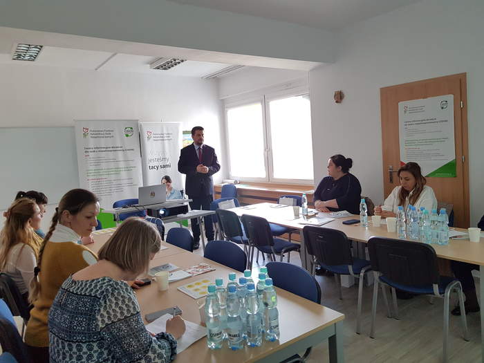 Pokaż zdjęcie: Sala, gdzie siedzą pracownicy OPS, w tle stoi Dyrektor Oddziału i przemawia, są duże okna w sali, na stołach jest catering, notatniki itp.