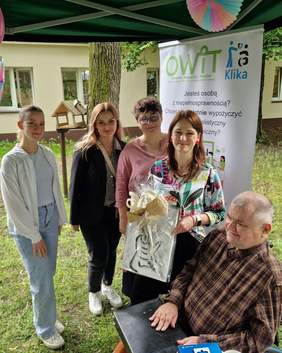 Pokaż zdjęcie: Grupa kilku osób pozuje do zdjęcia. Wśród nich mężczyzna na wózku. Obok kobieta z ozdobnie zapakowanym obrazem. W tle baner z logo OWIT