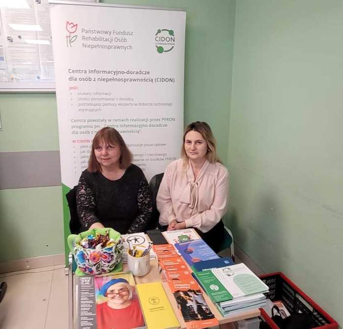 Pokaż zdjęcie: Stoisko informacyjne PFRON w placówce ZUS – dwie osoby siedzą przy stoliku z materiałami i ulotkami. W tle roll-up CIDON. Punkt udzielania porad dla osób z niepełnosprawnościami podczas dyżuru eksperckiego.
