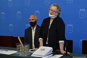 Pokaż zdjęcie: Za stołem  na którym leża materiały promocyjne stoi kobieta w siwych włosach, okularach ubrana w długą ciemną marynarkę. Za nią siedzi mężczyzna ubrany w biała koszulę i ciemną marynarkę  z maską na ustach.