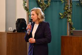 Pokaż zdjęcie: Zdjęcie kobiety blondynki ubranej w ciemny garnitur, kobieta się lekko uśmiecha, w ręku trzyma mikrofon, zdjęcie do połowy ciała.