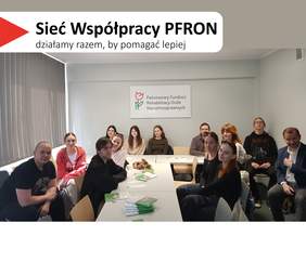 Pokaż zdjęcie: Kilkanaście osób siedzi przy długim białym stole z rozłożonymi ulotkami. Na ścianie za uczestnikami wisi tablica z napisem „Państwowy Fundusz Rehabilitacji Osób Niepełnosprawnych”. W lewym górnym rogu widnieje hasło „Sieć Współpracy PFRON – działamy razem, by pomagać lepiej”. Po lewej stronie zdjęcia widać okno z pionowymi żaluzjami.