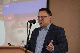 Pokaż zdjęcie: Przy mównicy stoi mężczyzna w garniturze, gestykuluje rękami