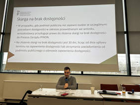 Pokaż zdjęcie: mężczyzna siedzący za stołem prezydialnym w Sali konferencyjnej, na ekranie wyświetla się prezentacja