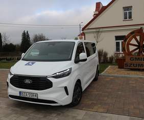 Pokaż zdjęcie: Biały samochód marki Ford przystosowany do przewozu osób z niepełnosprawnościami, zaparkowany przed budynkiem Gminy Szumowo.
