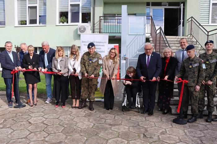 Pokaż zdjęcie: Uroczyste przecięcie czerwonej wstęgi przed budynkiem Szkoły Rolniczej. Na pierwszym planie, na brukowanym placu, stoi w półkolu kilkanaście osób. Wśród nich znajdują się osoby w strojach biznesowych oraz uczniowie z klasy mundurowej. Wszyscy trzymają w rękach nożyczki i przecinają wstęgę. W tle widoczne są szerokie schody wejściowe oraz drzwi i okna budynku.