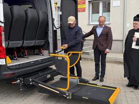 Pokaż zdjęcie: Mężczyzna obsługuje windę dla osób z niepełnosprawnościami w autobusie. Obok stoją dwaj mężczyźni przed budynkiem Urzędu Gminy Linia.