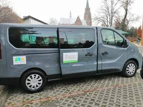 Pokaż zdjęcie: Na wypuzlowanym parkingu stoi duży szary bus z boku pojazdu naklejka z logo i napis powiat raciborski oraz logo i napis dofinansowano ze środków PFRON