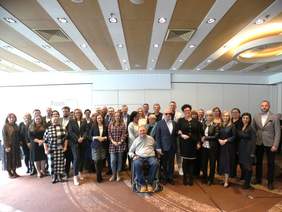 Pokaż zdjęcie: W sali konferencyjnej pozuje do zdjęcia około 30 osób. Są to członkowie Forum Dialogu. Większość osób się uśmiecha. W pierwszym rzędzie siedzi pan na wózku inwalidzkim. 