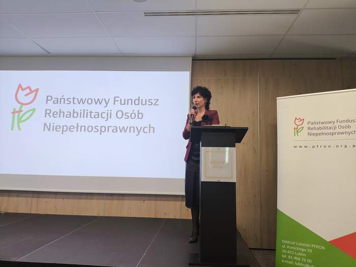 Pokaż zdjęcie: Przy mównicy stoi kobieta, w tle prezentacja z logo PFRON.