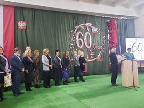 Pokaż zdjęcie: Na środku sali stoją nauczyciele i nauczycielki, którzy składają życzenia i gratulacje dyrekcji ośrodka. Kobieta, która stoi przy mównicy, trzyma w dłoniach piękny bukiet kolorowych kwiatów. 