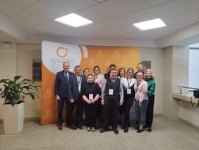 Zdęcie grupowe kilkunastu osób, mężczyzn i kobiet, stoją, pozują, uśmiechają się. Jasna sala, za nimi banery w kolorystyce biało-pomarańczowej