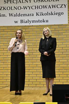 Pokaż zdjęcie: Dwie kobiety stojące na scenie na tle żółtej ceglanej ściany. Jedna z nich trzyma mikrofon i przemawia, ubrana jest w jasną bluzkę i czarne spodnie. Druga kobieta stoi obok, ubrana w ciemny kostium, i słucha. Nad nimi widnieje napis informujący, że wydarzenie odbywa się w Specjalnym Ośrodku Szkolno-Wychowawczym im. Waldemara Kikolskiego w Białymstoku.