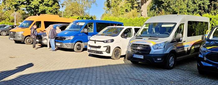 Pokaż zdjęcie: Zaparkowane busy stoją obok siebie na brukowanym parkingu w słoneczny dzień. Na szarym i niebieskim pojeździe widoczne są oznaczenia Fundacji oraz napisy „USŁUGI TRANSPORTOWE”. W tle drzewa i zieleń