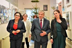 Pokaż zdjęcie: Fragment korytarza którym idą ludzie. Na pierwszym planie dwie kobiety i mężczyzna elegancko ubrani oglądają wnętrze placówki i rozmawiają.