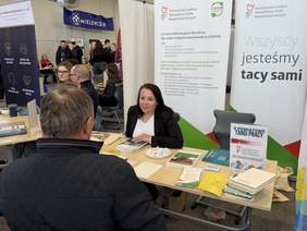 Pokaż zdjęcie: Stoisko informacyjne na targach pracy. Kobieta w marynarce rozmawia z mężczyzną siedzącym naprzeciwko przy stole z ulotkami i materiałami. W tle inne stoiska, roll-upy i uczestnicy wydarzenia.