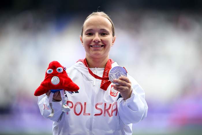 Pokaż zdjęcie: Kobieta trzyma srebny medal, jest ubrana w biały dres i uśmiechnięta