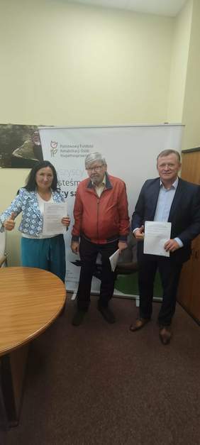 Pokaż zdjęcie: W gabinecie dyrektora Łódzkiego Oddziału PFRON stoi kobieta i dwóch mężczyzn. Zawarli umowy o dofinansowania. W tle widać baner z logo PFRON. 