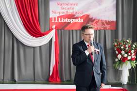 Pokaż zdjęcie: Mężczyzna w garniturze przemawia do mikrofonu podczas uroczystości z okazji Narodowego Święta Niepodległości. W tle widoczna jest biało-czerwona dekoracja i baner z napisem „11 Listopada”.