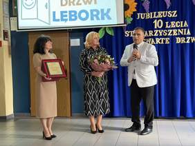 Pokaż zdjęcie: Trzy osoby podczas jubileuszu 10-lecia Stowarzyszenia Otwarte Drzwi w Lęborku; kobieta w beżowej sukience trzyma tablicę z wyróżnieniem, druga z bukietem kwiatów, a mężczyzna w jasnej marynarce przemawia z mikrofonem na tle niebieskiej kurtyny.