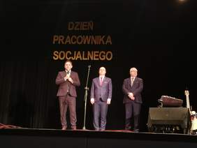 Pokaż zdjęcie: Na scenie Grajewskiego Centrum Kultury stoi trzech mężczyzn, pierwszy z nich, licząc od lewej strony, trzyma w rękach mikrofon i przemawia. Nad nimi umieszczony jest napis Dzień Pracownika Socjalnego.