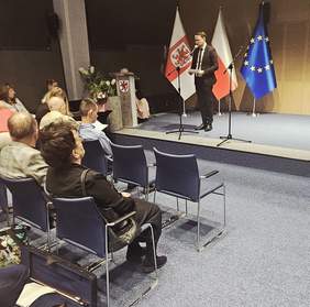 Pokaż zdjęcie: Szeroki kadr przedstawiający prelegenta lub mówcę stojącego na podwyższonej scenie oraz fragment widowni. Mężczyzna ubrany w ciemny garnitur i krawat stoi na środku sceny, trzymając coś w ręku (prawdopodobnie notatki lub nagrodę/upominek) i przemawia do mikrofonów. Za nim widoczne są te same trzy flagi co na pierwszym zdjęciu: flaga z herbem województwa zachodniopomorskiego (czerwony gryf), flaga Polski i flaga Unii Europejskiej. Scena jest dobrze oświetlona, tło ma jasnobrązową, panelową teksturę. Na pierwszym planie widać kilkunastu widzów siedzących na niebieskich krzesłach, na niebieskiej wykładzinie. Po lewej stronie w dolnym rogu na podłodze leży otwarty futerał na instrument smyczkowy, prawdopodobnie skrzypce lub altówkę.