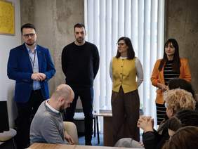Pokaż zdjęcie: Cztery osoby stojące obok siebie prowadzą prezentację dla uczestników spotkania. Przed nimi siedzą osoby przy stole konferencyjnym. W tle znajdują się pionowe rolety okienne i ściana w stylu industrialnym.