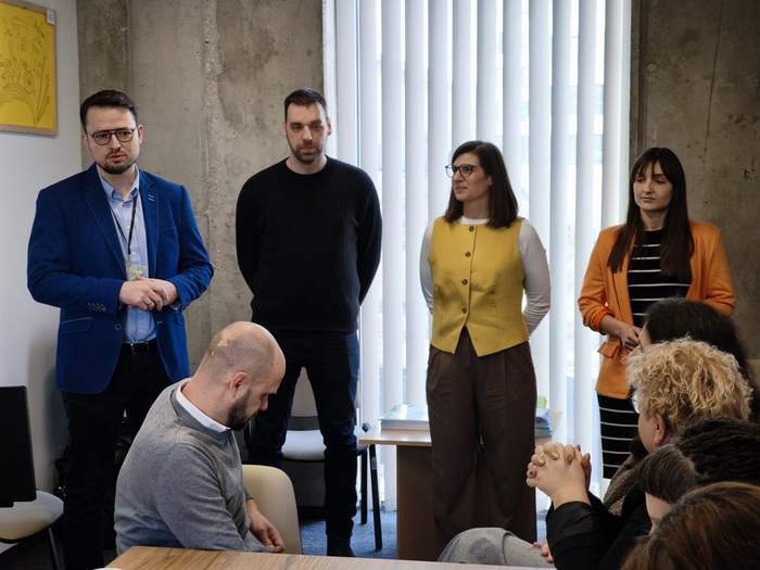 Pokaż zdjęcie: Cztery osoby stojące obok siebie prowadzą prezentację dla uczestników spotkania. Przed nimi siedzą osoby przy stole konferencyjnym. W tle znajdują się pionowe rolety okienne i ściana w stylu industrialnym.